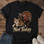 Black Gildan 64000 unisex Thanksgiving cat T-shirt laid flat on wooden planks with rolled short sleeves, folded blue jeans and light gray lace-up sneakers. The vintage chest artwork shows a startled brown tabby running from a turkey with raised wings and a fanned orange-brown tail. Large cream "Not Today" lettering beneath the animals stands out against the black fabric.