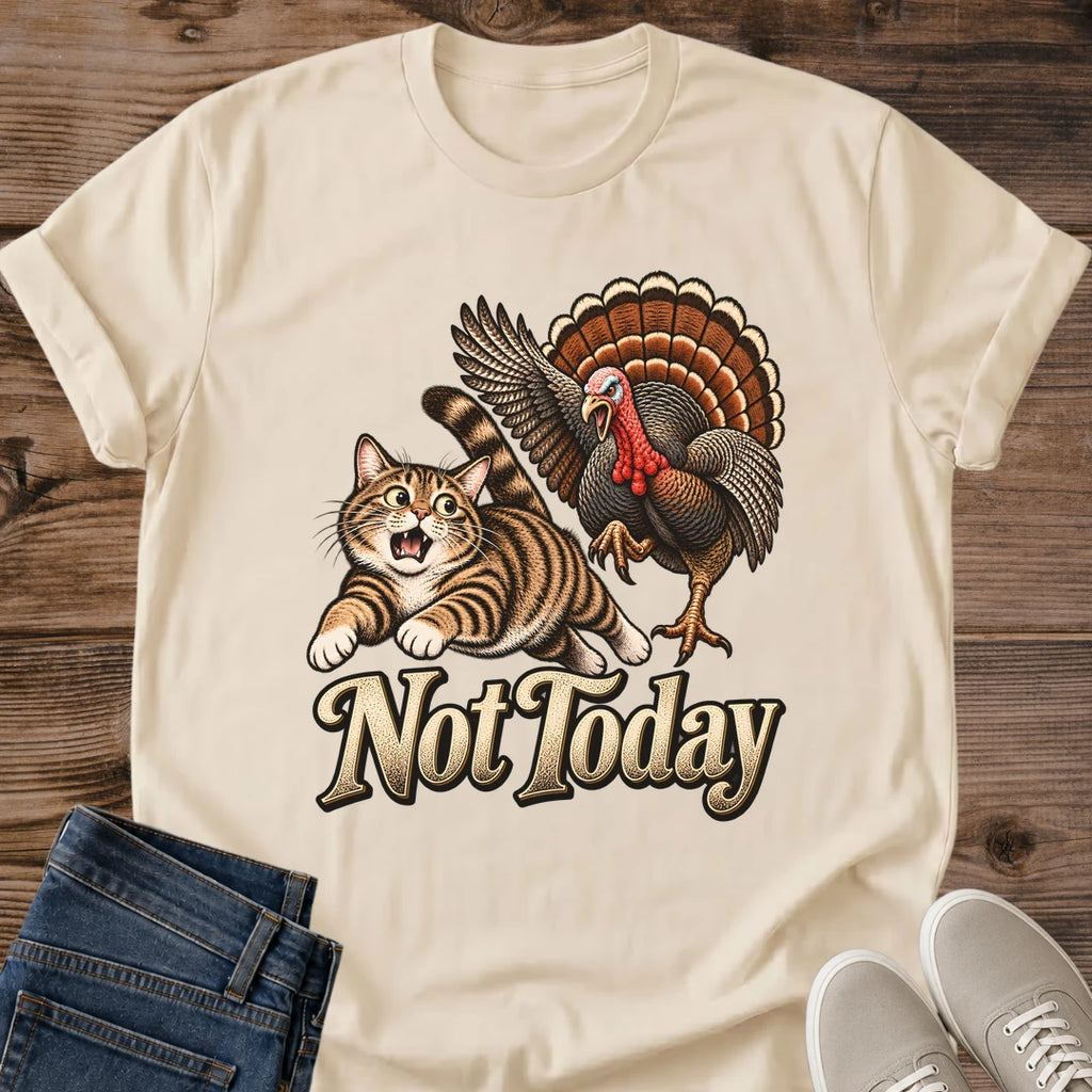 Sand beige Gildan 64000 unisex Thanksgiving cat T-shirt shown from the front, laid flat on rustic wood with rolled short sleeves, folded blue jeans and light gray lace-up sneakers. The vintage chest graphic shows a wide-eyed brown tabby running from a turkey with raised wings and a fanned brown and orange tail, above cream "Not Today" lettering with a dark outline.