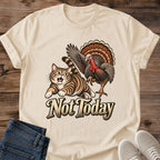 Sand beige Gildan 64000 unisex Thanksgiving cat T-shirt shown from the front, laid flat on rustic wood with rolled short sleeves, folded blue jeans and light gray lace-up sneakers. The vintage chest graphic shows a wide-eyed brown tabby running from a turkey with raised wings and a fanned brown and orange tail, above cream "Not Today" lettering with a dark outline.