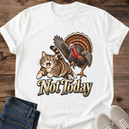 White Gildan 64000 unisex Thanksgiving cat T-shirt shown from the front on rustic wood, with a crew neck and rolled short sleeves. A detailed vintage graphic depicts a wide-eyed brown striped tabby running from a turkey with raised wings, a red wattle and a fan of brown and orange feathers. Cream "Not Today" lettering has a dark outline; folded blue jeans and light gray sneakers sit below.