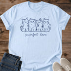 Light Blue Gildan 64000 unisex Purrfect Love Four Cats T-shirt laid flat on wooden planks beside folded dark blue jeans and light gray lace-up sneakers. Navy line art shows four cats sitting closely together, with striped, fluffy, patch-marked and smooth coats, above lowercase "purrfect love" script. The pastel blue shirt has a round crew neckline and rolled short sleeves, with the small cat illustration centered across the chest.