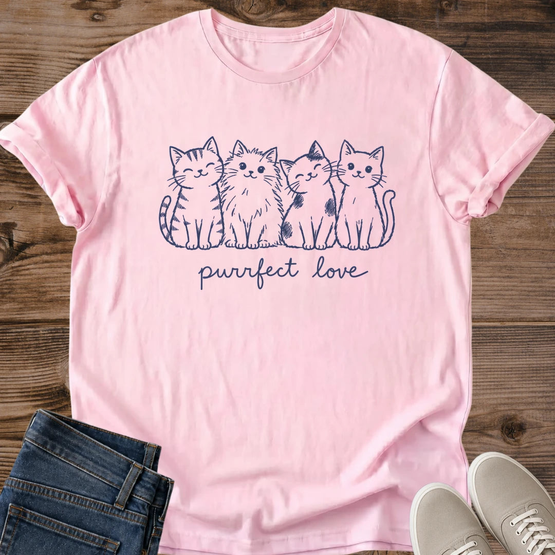Light Pink Gildan 64000 unisex Purrfect Love cat T-shirt in a front-view flat lay on wooden planks with folded blue jeans and light gray lace-up sneakers. The navy line-art chest print features four seated cats: a striped tabby, a fluffy cat, a patch-marked cat and a smooth-coated cat with a curved tail. Handwritten "purrfect love" lettering runs beneath them. The pale pink crewneck has rolled short sleeves, visible fabric folds and a stitched bottom hem.