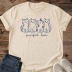 Sand beige Gildan 64000 unisex Purrfect Love Four Cats T-shirt laid flat on rustic wooden planks with rolled short sleeves and a crew neck. A centered navy line-art illustration shows four cats sitting shoulder to shoulder, their striped, fluffy, patch-marked and smooth coats creating distinct silhouettes. Lowercase "purrfect love" script sits underneath. Folded dark blue jeans overlap the lower left edge, with light gray sneakers at the lower right.
