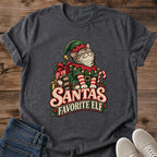 Dark Heather charcoal gray Gildan 64000 unisex Christmas cat T-shirt with rolled short sleeves and a crewneck. A tabby with closed eyes wears a green elf hat and outfit with red trim, gold bells, a buckled belt and red-and-white striped sleeves. Wrapped gifts, a candy cane and holly surround the cat above cream-and-red "Santa's Favorite Elf" lettering. Front flat lay on brown wood planks, with dark denim jeans at lower left and light gray lace-up sneakers at lower right.