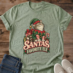 Heather Military Green with a mottled green texture Gildan 64000 unisex Christmas cat T-shirt with rolled short sleeves and a crewneck. A tabby with closed eyes wears a green elf hat and outfit with red trim, gold bells, a buckled belt and red-and-white striped sleeves. Wrapped gifts, a candy cane and holly surround the cat above cream-and-red "Santa's Favorite Elf" lettering. Front flat lay on brown wood planks, with dark denim jeans at lower left and light gray lace-up sneakers at lower right.