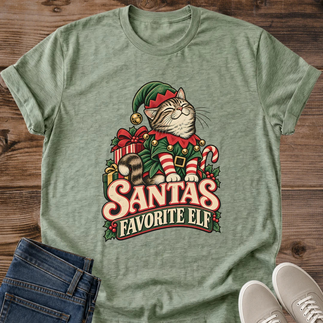 Heather Military Green with a mottled green texture Gildan 64000 unisex Christmas cat T-shirt with rolled short sleeves and a crewneck. A tabby with closed eyes wears a green elf hat and outfit with red trim, gold bells, a buckled belt and red-and-white striped sleeves. Wrapped gifts, a candy cane and holly surround the cat above cream-and-red "Santa's Favorite Elf" lettering. Front flat lay on brown wood planks, with dark denim jeans at lower left and light gray lace-up sneakers at lower right.