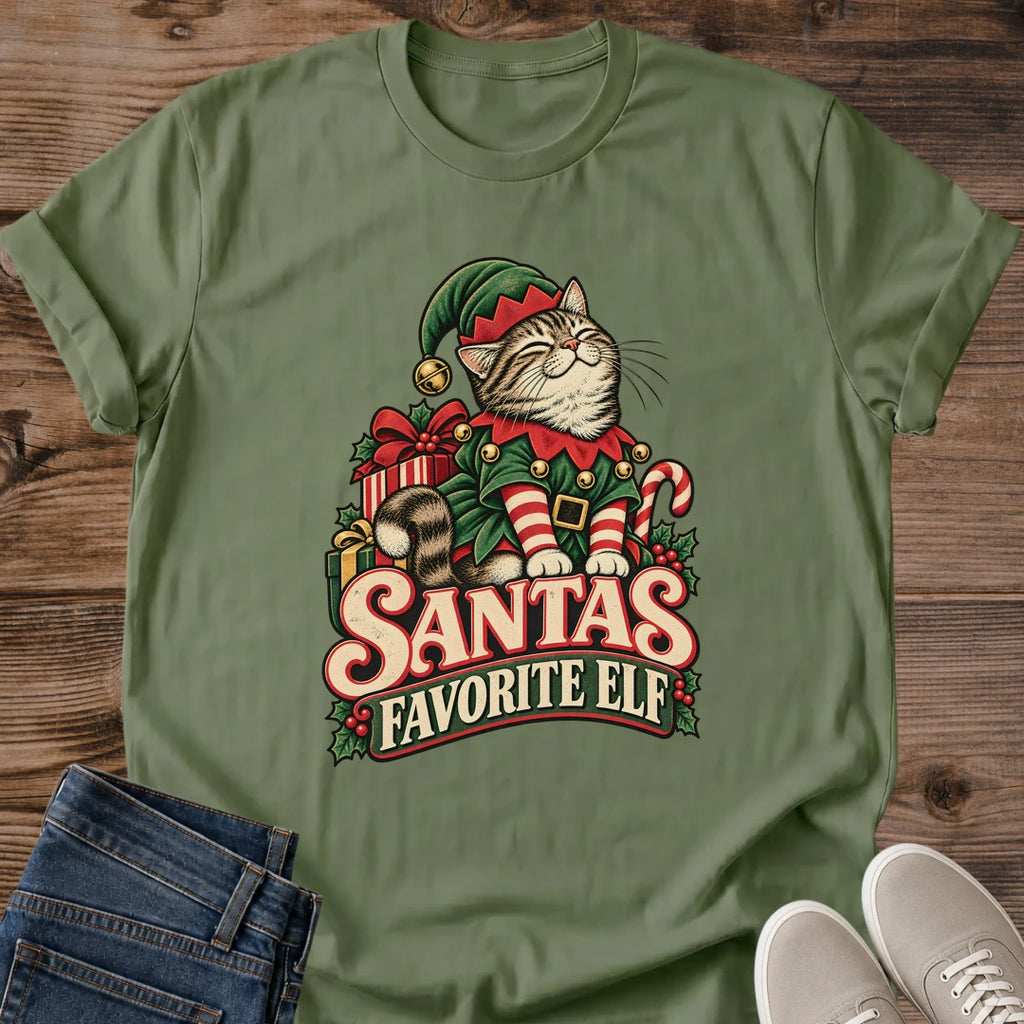 Military Green olive Gildan 64000 unisex Christmas cat T-shirt with rolled short sleeves and a crewneck. A tabby with closed eyes wears a green elf hat and outfit with red trim, gold bells, a buckled belt and red-and-white striped sleeves. Wrapped gifts, a candy cane and holly surround the cat above cream-and-red "Santa's Favorite Elf" lettering. Front flat lay on brown wood planks, with dark denim jeans at lower left and light gray lace-up sneakers at lower right.