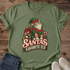 Military Green olive Gildan 64000 unisex Christmas cat T-shirt with rolled short sleeves and a crewneck. A tabby with closed eyes wears a green elf hat and outfit with red trim, gold bells, a buckled belt and red-and-white striped sleeves. Wrapped gifts, a candy cane and holly surround the cat above cream-and-red "Santa's Favorite Elf" lettering. Front flat lay on brown wood planks, with dark denim jeans at lower left and light gray lace-up sneakers at lower right.