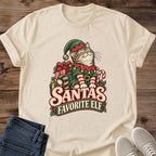 Sand beige Gildan 64000 unisex Christmas cat T-shirt with rolled short sleeves and a crewneck. A tabby with closed eyes wears a green elf hat and outfit with red trim, gold bells, a buckled belt and red-and-white striped sleeves. Wrapped gifts, a candy cane and holly surround the cat above cream-and-red "Santa's Favorite Elf" lettering. Front flat lay on brown wood planks, with dark denim jeans at lower left and light gray lace-up sneakers at lower right.