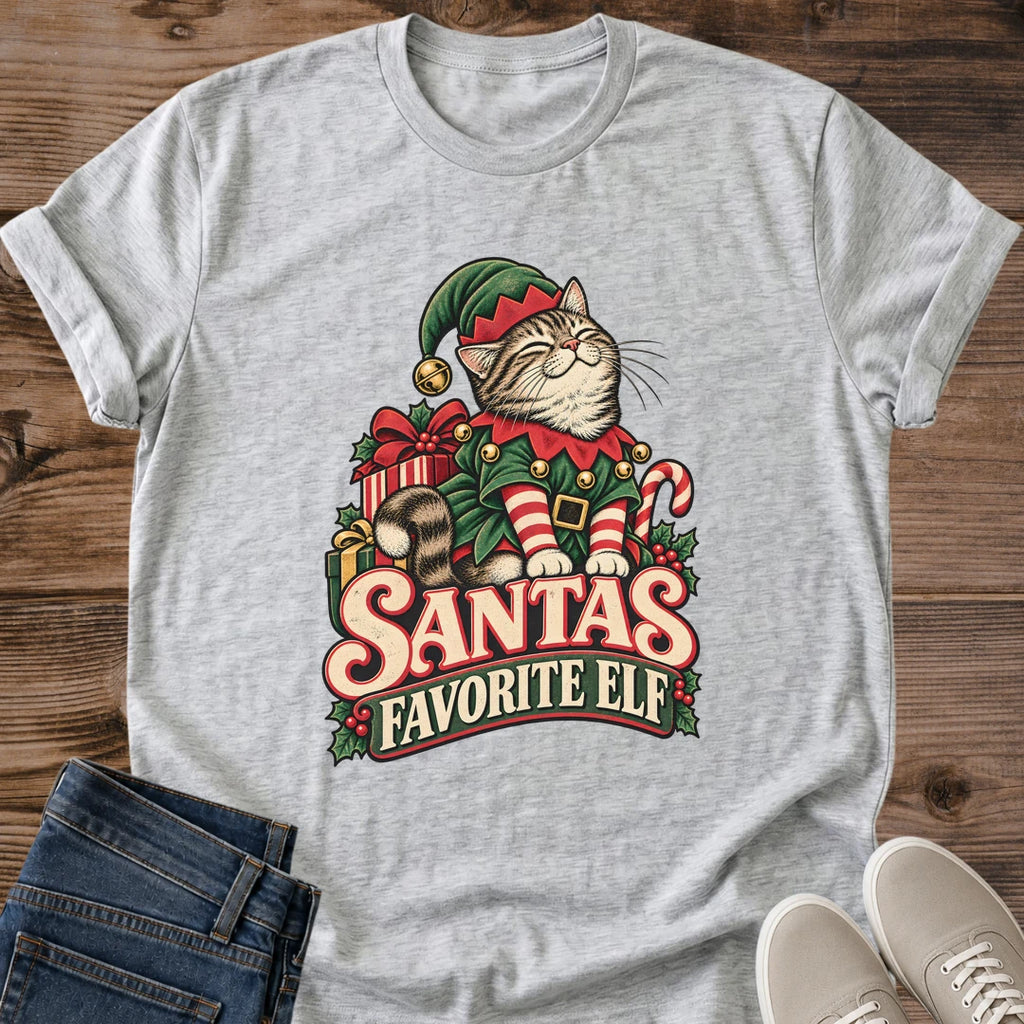 Sport Grey light heather gray Gildan 64000 unisex Christmas cat T-shirt with rolled short sleeves and a crewneck. A tabby with closed eyes wears a green elf hat and outfit with red trim, gold bells, a buckled belt and red-and-white striped sleeves. Wrapped gifts, a candy cane and holly surround the cat above cream-and-red "Santa's Favorite Elf" lettering. Front flat lay on brown wood planks, with dark denim jeans at lower left and light gray lace-up sneakers at lower right.
