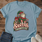 Stone Blue muted blue Gildan 64000 unisex Christmas cat T-shirt with rolled short sleeves and a crewneck. A tabby with closed eyes wears a green elf hat and outfit with red trim, gold bells, a buckled belt and red-and-white striped sleeves. Wrapped gifts, a candy cane and holly surround the cat above cream-and-red "Santa's Favorite Elf" lettering. Front flat lay on brown wood planks, with dark denim jeans at lower left and light gray lace-up sneakers at lower right.
