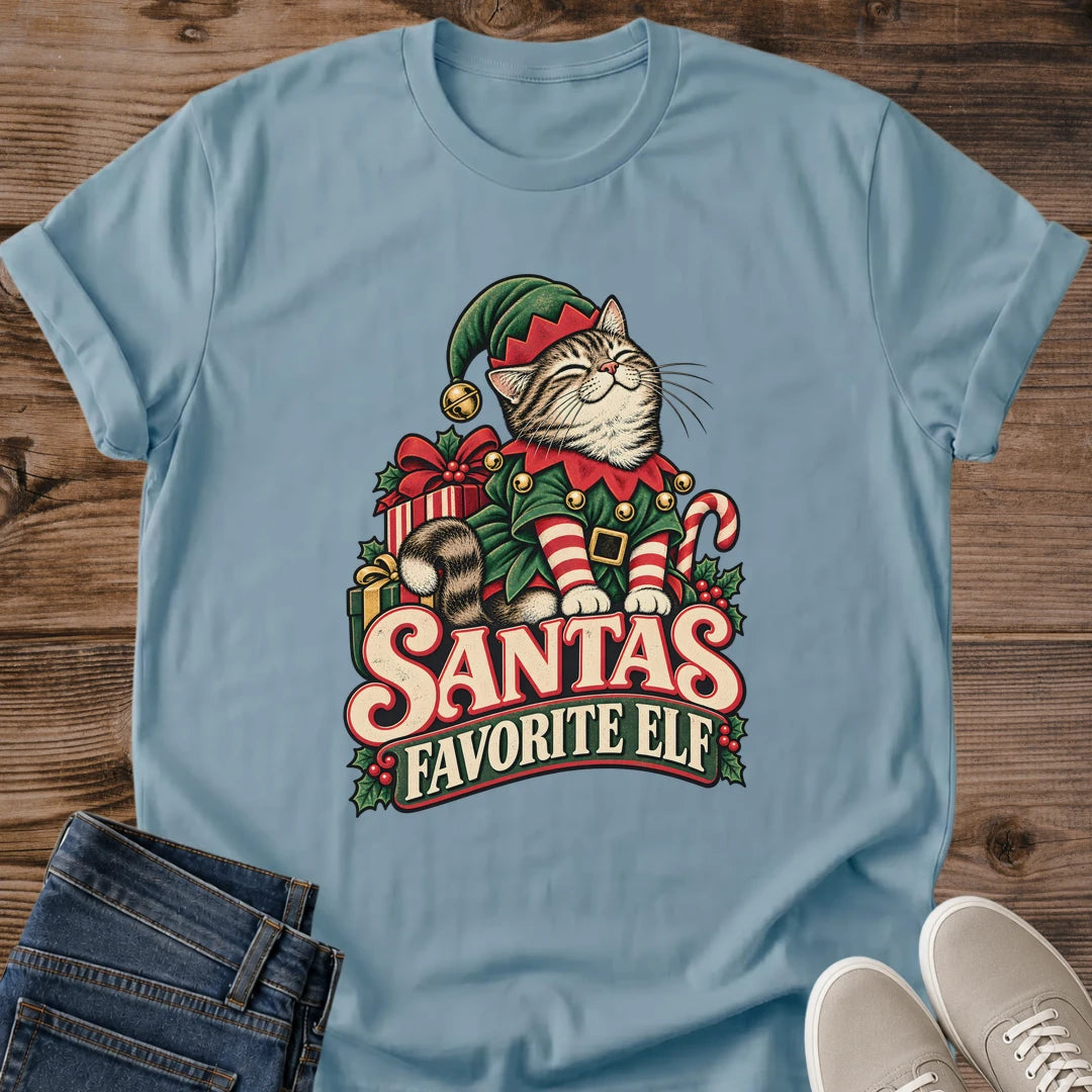 Stone Blue muted blue Gildan 64000 unisex Christmas cat T-shirt with rolled short sleeves and a crewneck. A tabby with closed eyes wears a green elf hat and outfit with red trim, gold bells, a buckled belt and red-and-white striped sleeves. Wrapped gifts, a candy cane and holly surround the cat above cream-and-red "Santa's Favorite Elf" lettering. Front flat lay on brown wood planks, with dark denim jeans at lower left and light gray lace-up sneakers at lower right.