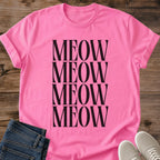 Azalea pink Gildan 64000 unisex Stacked Meow cat T-shirt shown from the front on rustic wooden planks. The bright pink crewneck tee features four stacked rows of "MEOW" in tall black uppercase serif lettering, forming a large minimalist cat typography print. Rolled short sleeves frame the shirt, with folded dark blue jeans at the lower left and light gray lace-up sneakers at the lower right.