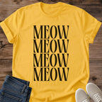 Daisy yellow Gildan 64000 unisex Stacked Meow cat T-shirt in a front-view flat lay on wooden planks. Four rows of tall black uppercase serif letters spell "MEOW" down the center, creating a bold rectangular typography design against the bright yellow fabric. The crewneck shirt has rolled short sleeves, with folded dark blue jeans and light gray lace-up sneakers framing the bottom.
