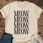 Sand beige Gildan 64000 unisex Stacked Meow cat T-shirt in a flat lay on rustic wooden planks beside folded dark blue jeans and light gray lace-up sneakers. Four closely stacked rows of "MEOW" in tall black uppercase serif letters form a rectangular print down the front. The light beige shirt is shown with a round crew neckline, rolled short sleeves and gentle folds near the hem.