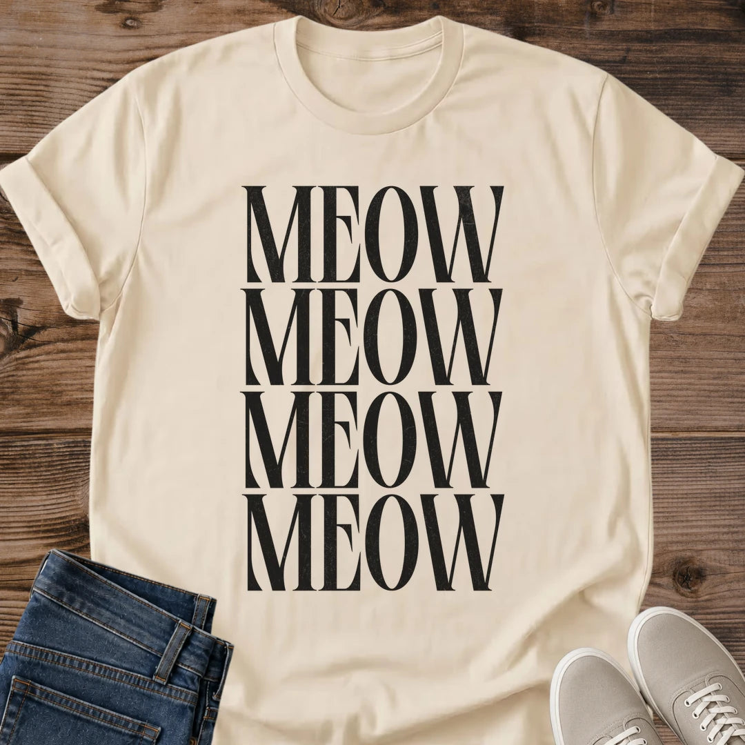Sand beige Gildan 64000 unisex Stacked Meow cat T-shirt in a flat lay on rustic wooden planks beside folded dark blue jeans and light gray lace-up sneakers. Four closely stacked rows of "MEOW" in tall black uppercase serif letters form a rectangular print down the front. The light beige shirt is shown with a round crew neckline, rolled short sleeves and gentle folds near the hem.
