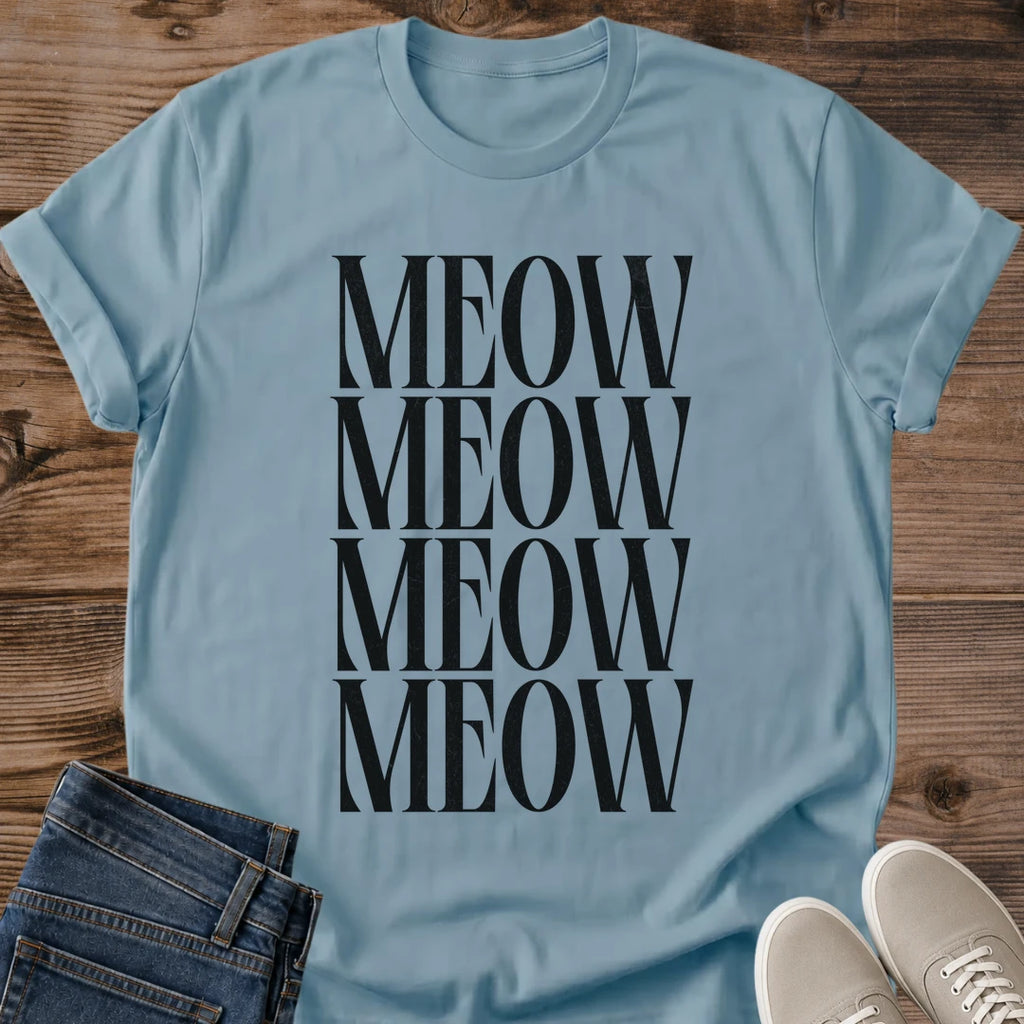 Stone Blue Gildan 64000 unisex Stacked Meow cat T-shirt laid flat on rustic wood, showing a crew neck and rolled short sleeves. The muted blue shirt has a centered typography design with "MEOW" repeated in four closely stacked rows of tall black uppercase serif letters. Folded dark blue jeans overlap the lower left edge, and light gray lace-up sneakers sit at the lower right.