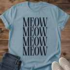 Stone Blue Gildan 64000 unisex Stacked Meow cat T-shirt laid flat on rustic wood, showing a crew neck and rolled short sleeves. The muted blue shirt has a centered typography design with "MEOW" repeated in four closely stacked rows of tall black uppercase serif letters. Folded dark blue jeans overlap the lower left edge, and light gray lace-up sneakers sit at the lower right.