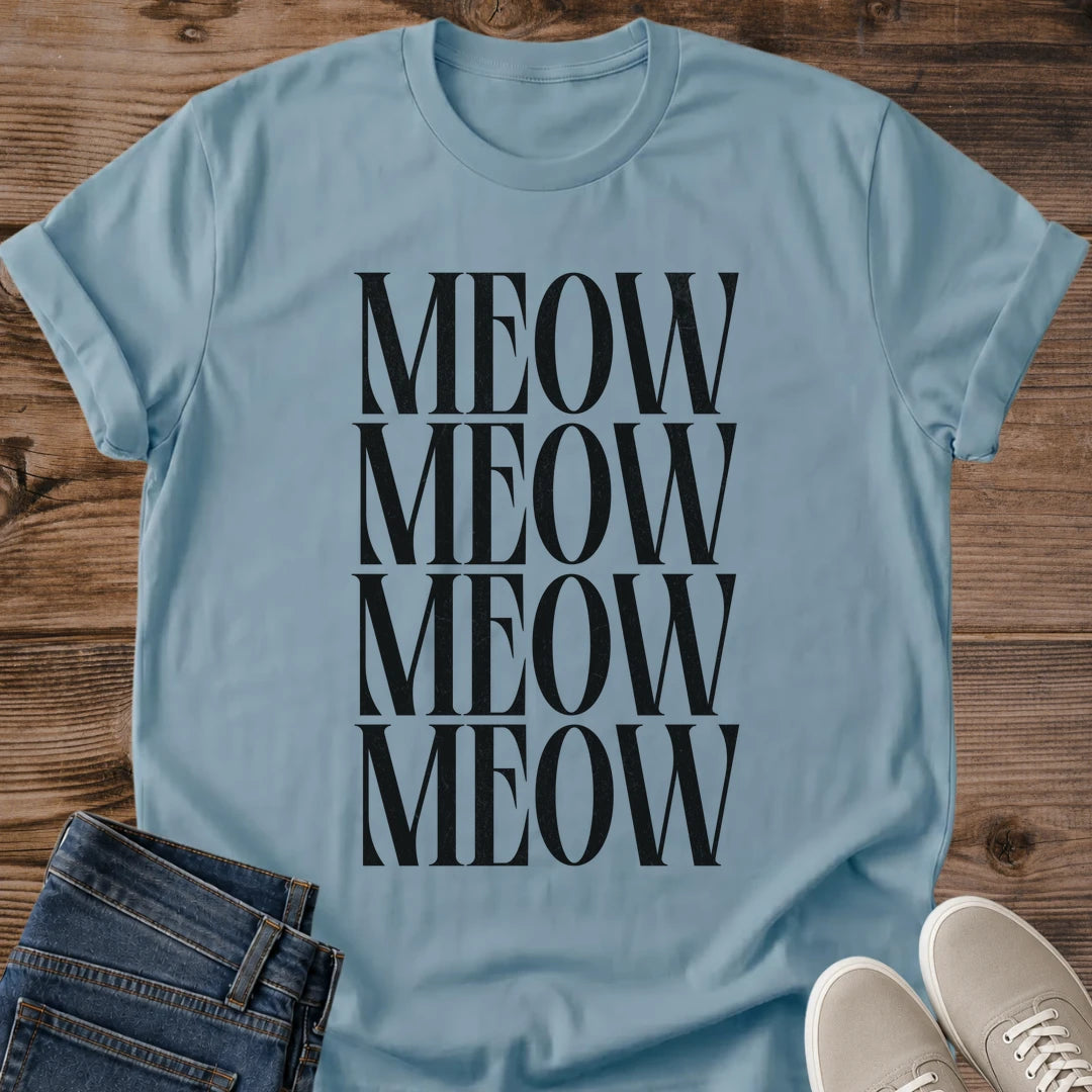 Stone Blue Gildan 64000 unisex Stacked Meow cat T-shirt laid flat on rustic wood, showing a crew neck and rolled short sleeves. The muted blue shirt has a centered typography design with "MEOW" repeated in four closely stacked rows of tall black uppercase serif letters. Folded dark blue jeans overlap the lower left edge, and light gray lace-up sneakers sit at the lower right.