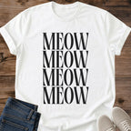 White Gildan 64000 unisex Stacked Meow cat T-shirt laid flat on a rustic wooden background, styled with folded dark blue jeans and light gray lace-up sneakers. The minimalist cat typography design repeats "MEOW" in four closely stacked rows of tall black uppercase serif lettering. The front view shows the round crew neckline, rolled short sleeves and black-on-white print.