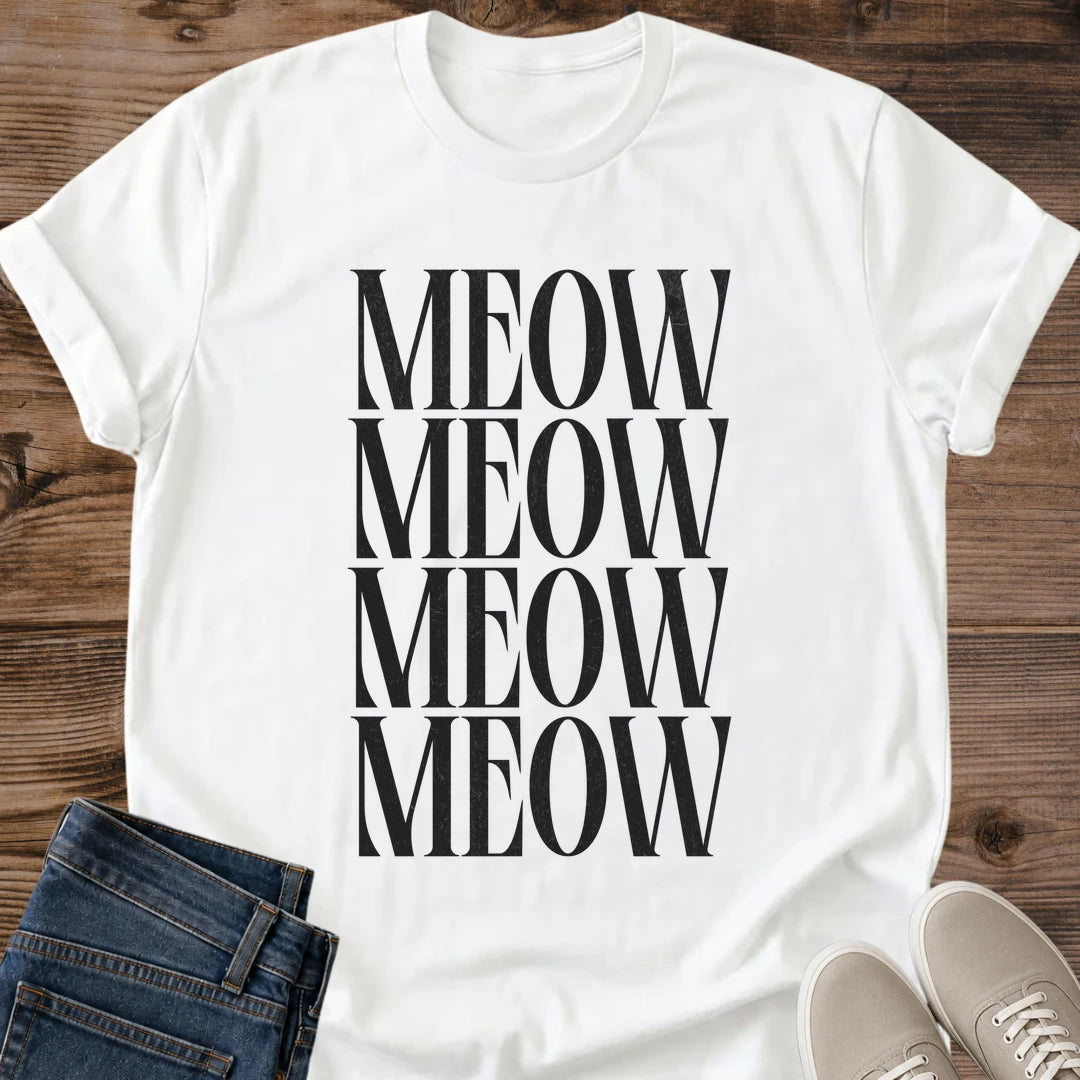 White Gildan 64000 unisex Stacked Meow cat T-shirt laid flat on a rustic wooden background, styled with folded dark blue jeans and light gray lace-up sneakers. The minimalist cat typography design repeats "MEOW" in four closely stacked rows of tall black uppercase serif lettering. The front view shows the round crew neckline, rolled short sleeves and black-on-white print.