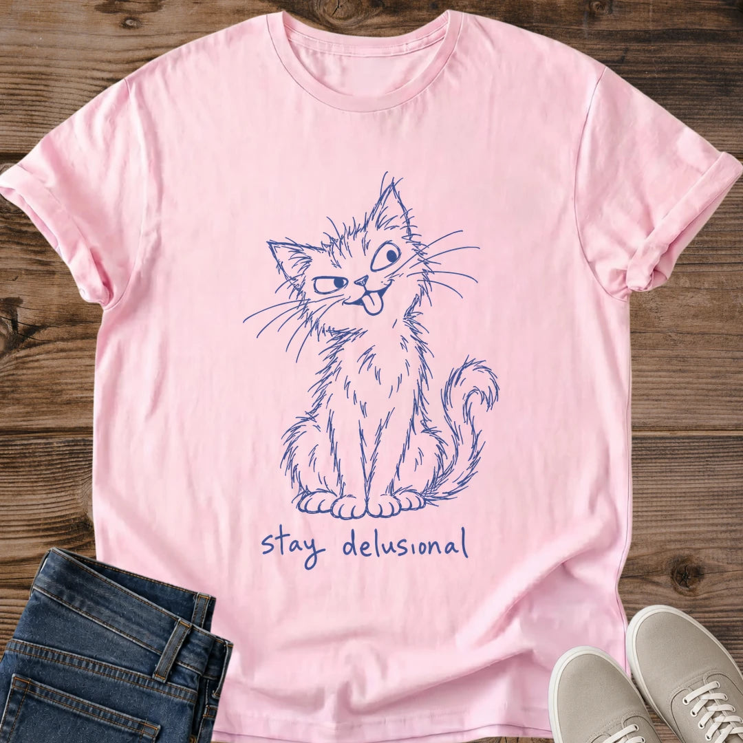 Light Pink Stay Delusional Cat Sketch T-Shirt, a Gildan 64000 unisex crewneck with rolled sleeves, soft folds and a visible stitched hem. Blue line art depicts a shaggy seated cat with uneven eyes, long whiskers, tongue sticking out and a curled tail beside its body. Handwritten "stay delusional" appears beneath the cat. Styled flat on brown wood planks with dark denim jeans at lower left and light gray lace-up sneakers at lower right.
