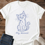 White Stay Delusional Cat Sketch T-Shirt, a Gildan 64000 unisex crewneck laid flat with rolled short sleeves. The navy blue drawing shows a seated shaggy cat with a tilted head, asymmetrical eyes, long whiskers, tongue sticking out and a curled tail on the right. Lowercase handwritten "stay delusional" appears beneath its paws. Rustic brown wood backdrop with folded dark blue jeans at lower left and light gray lace-up sneakers at lower right.